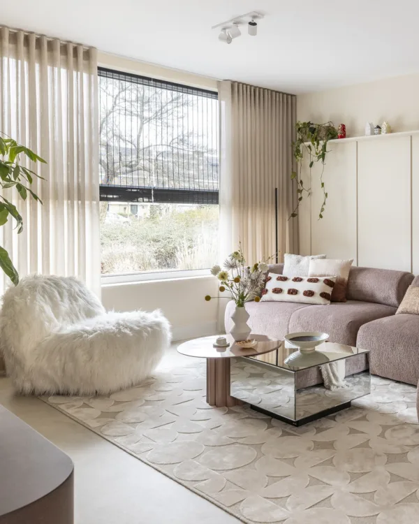 Woonkamer met betonlook vinylvloer en zacht vloerkleed, sfeervolle inbetween gordijnen en zwarte geweven hout vouwgordijnen.